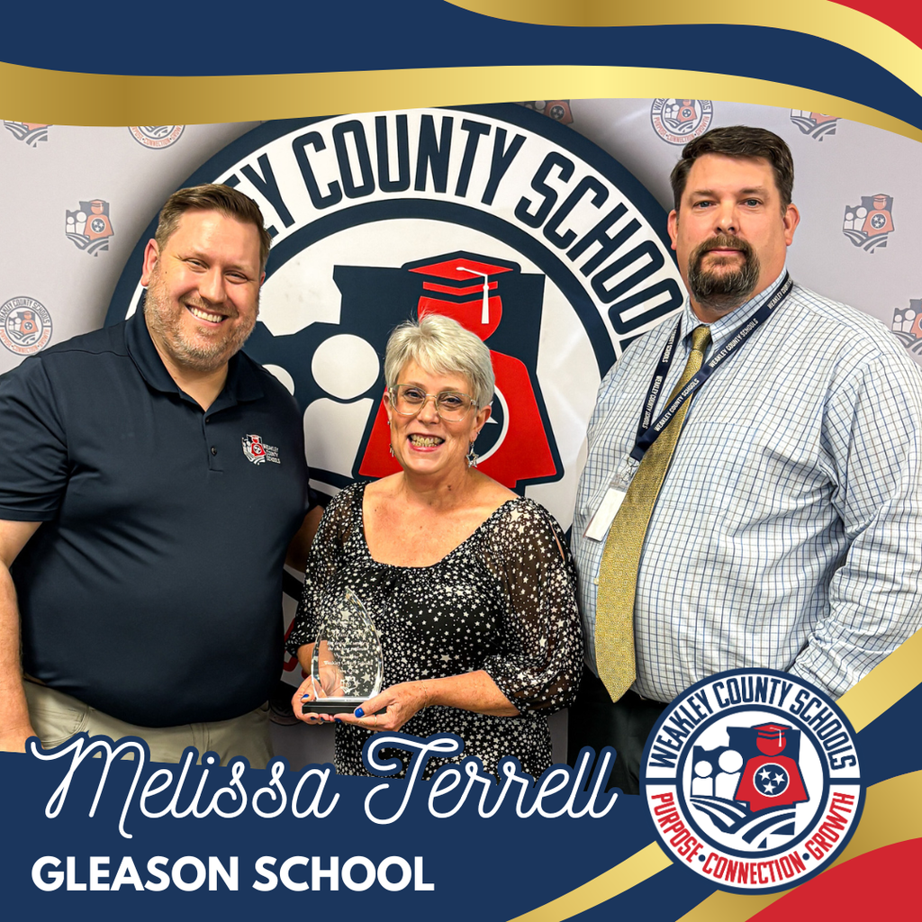 Three individuals stand together; a woman holds an award in her hands, smiling. The background features the logo for a school.