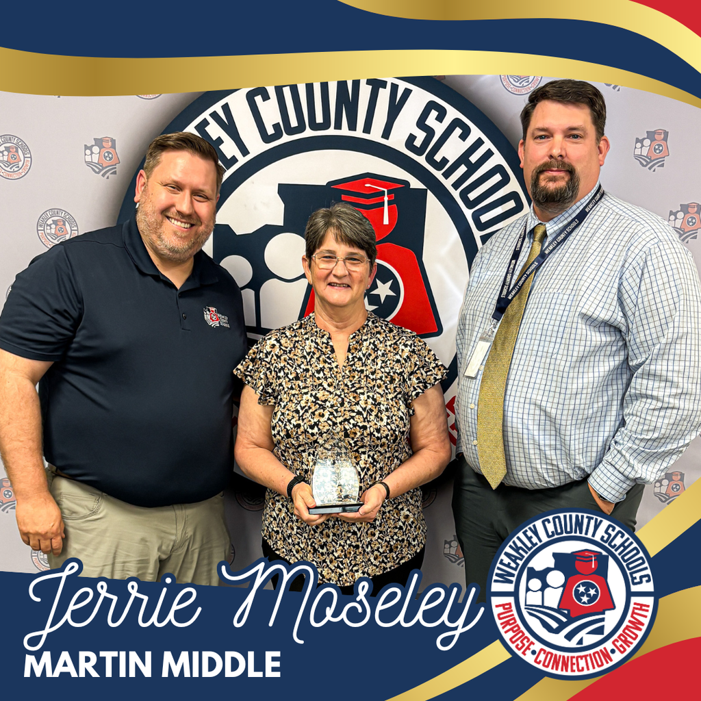 Three adults stand in front of a colorful backdrop with a school logo. The woman holds an award.