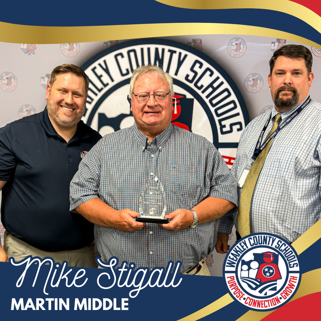 Three men pose for a photo with a school sign in the background. One holds a trophy.