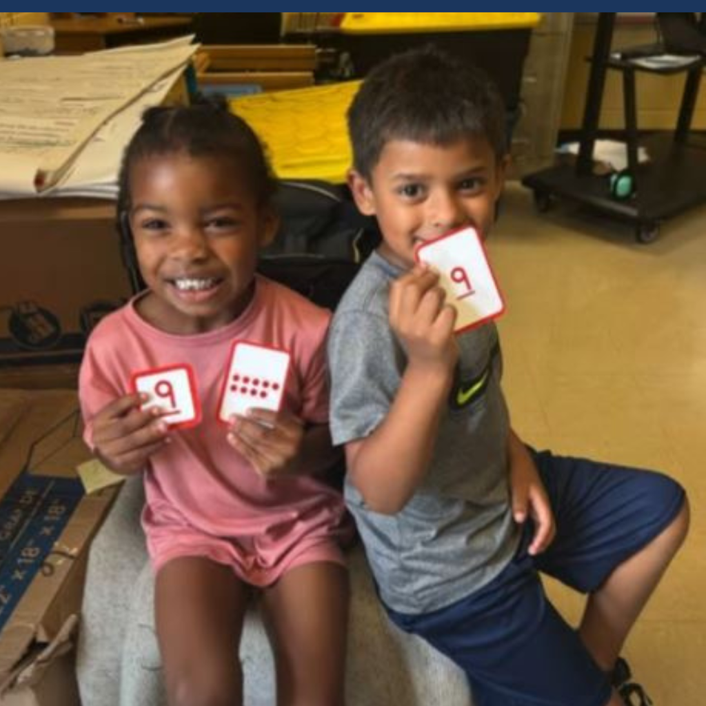 Two children in a classroom, one holding cards with numbers 9 and 9, the other holding a card with number 9.