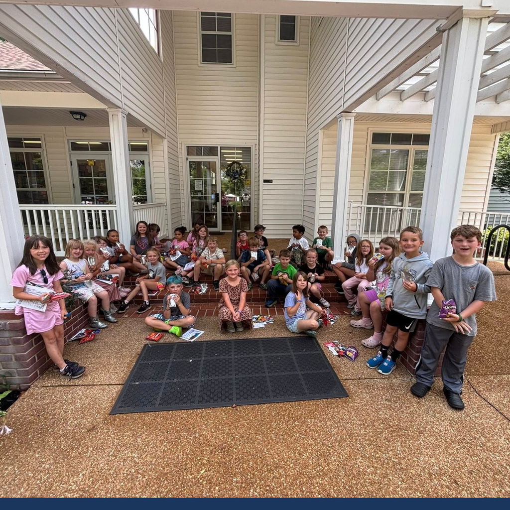 Group of children and two adults standing on a porch, holding books and other items, with a white building in the background.