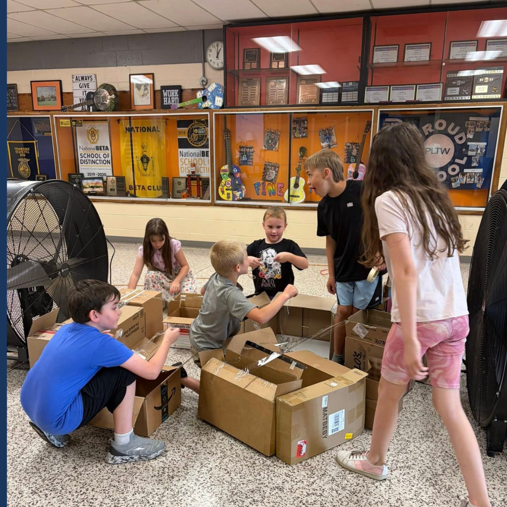 Children playing with cardboard boxes in a classroom with a clock and framed pictures on the wall.