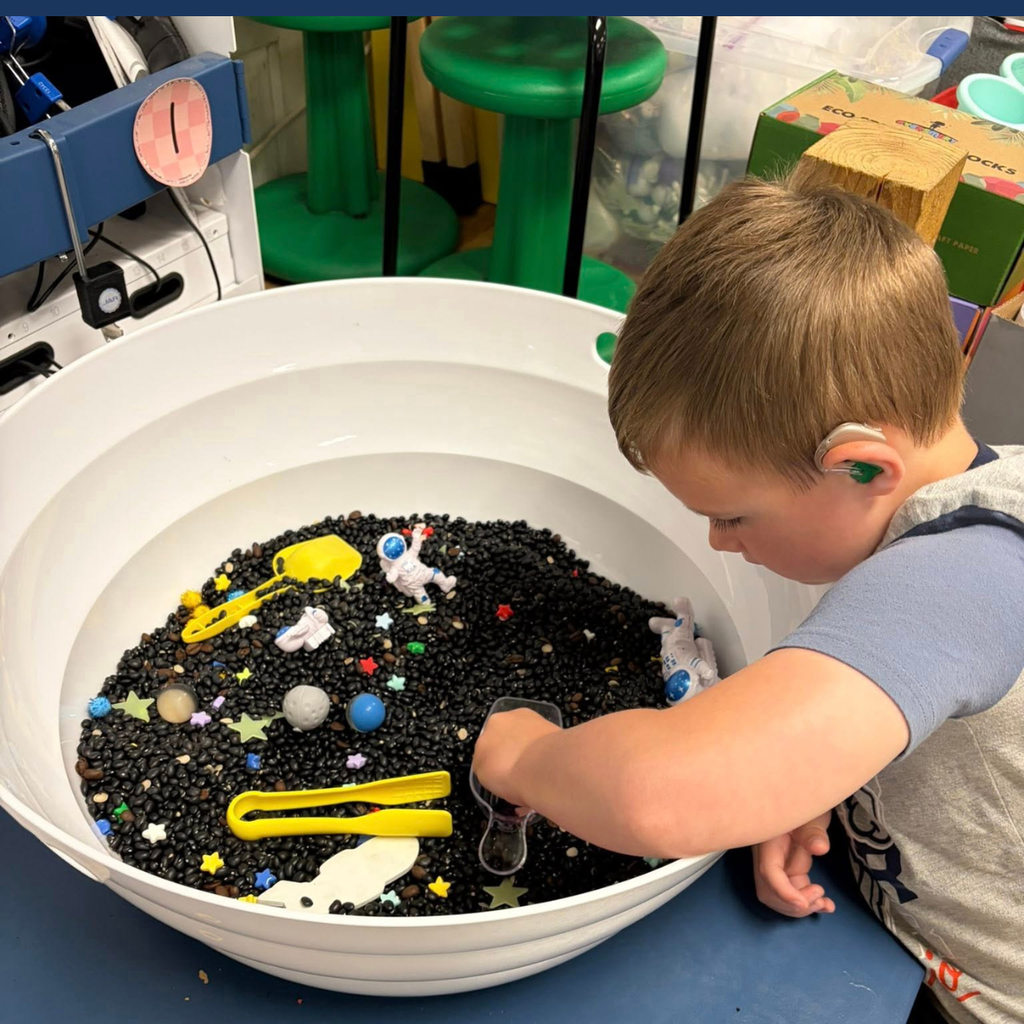 A child plays with toys in a large white sensory bin filled with black beads.