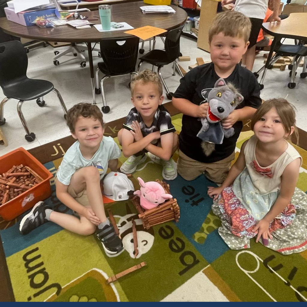 Four young children sitting in a classroom. Each child holds an item or is next to one.