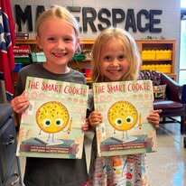 Two children holding books titled "The Smart Cookie" stand smiling in a classroom. Behind them, a wooden sign says "Masterspace."