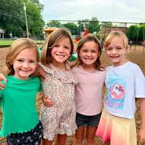 Four young girls stand together in a playground with a tree in the background. One girl has blonde hair, another has brown hair.