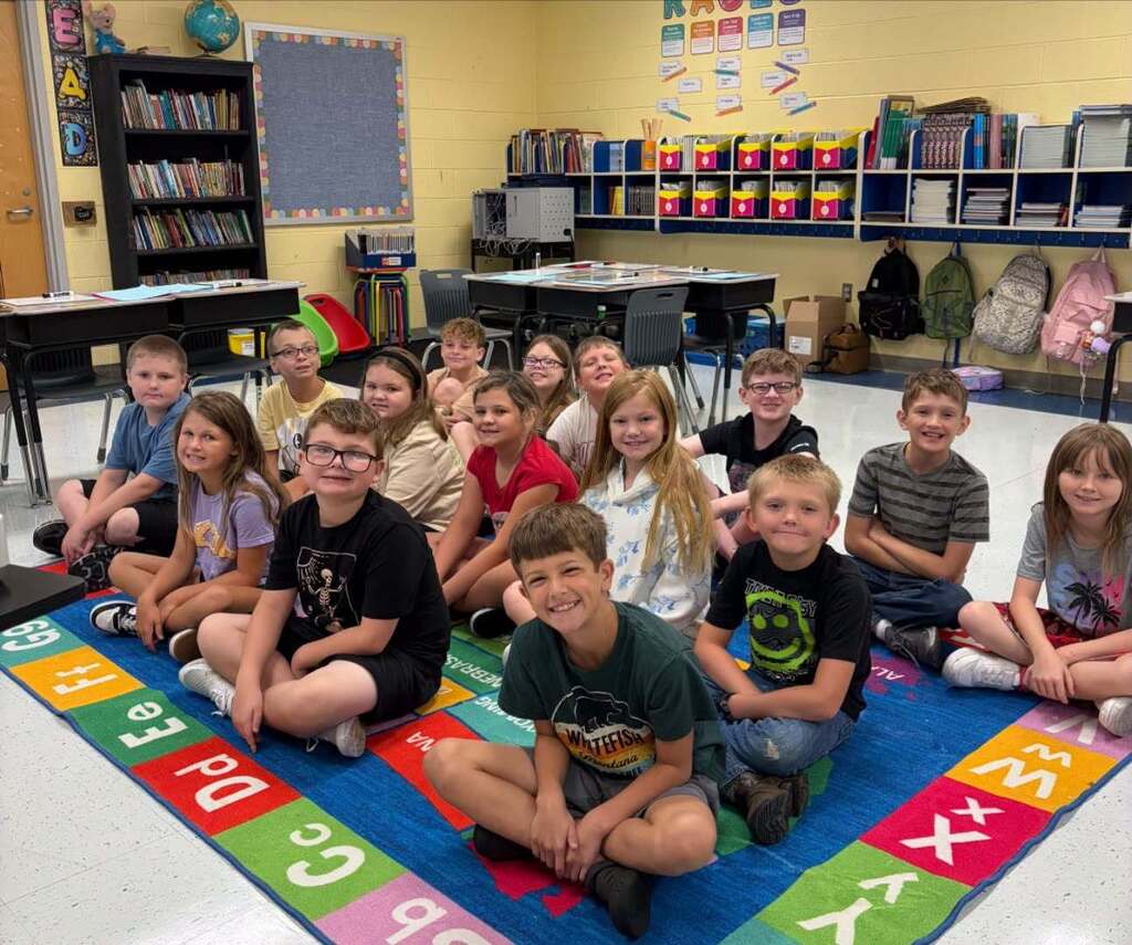 A classroom with children sitting in a circle on a colorful floor mat. Bookshelves and desks are visible in the background.