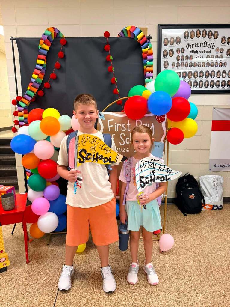 Two children stand with smiles in front of a colorful balloon arch and signs that read "First Day of School".