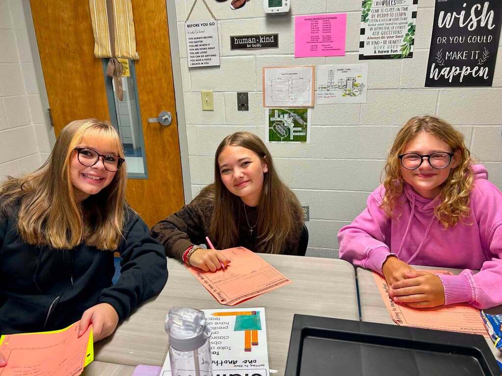 Three girls seated around a table in a classroom, each holding papers and a pen, with posters and a door in the background.