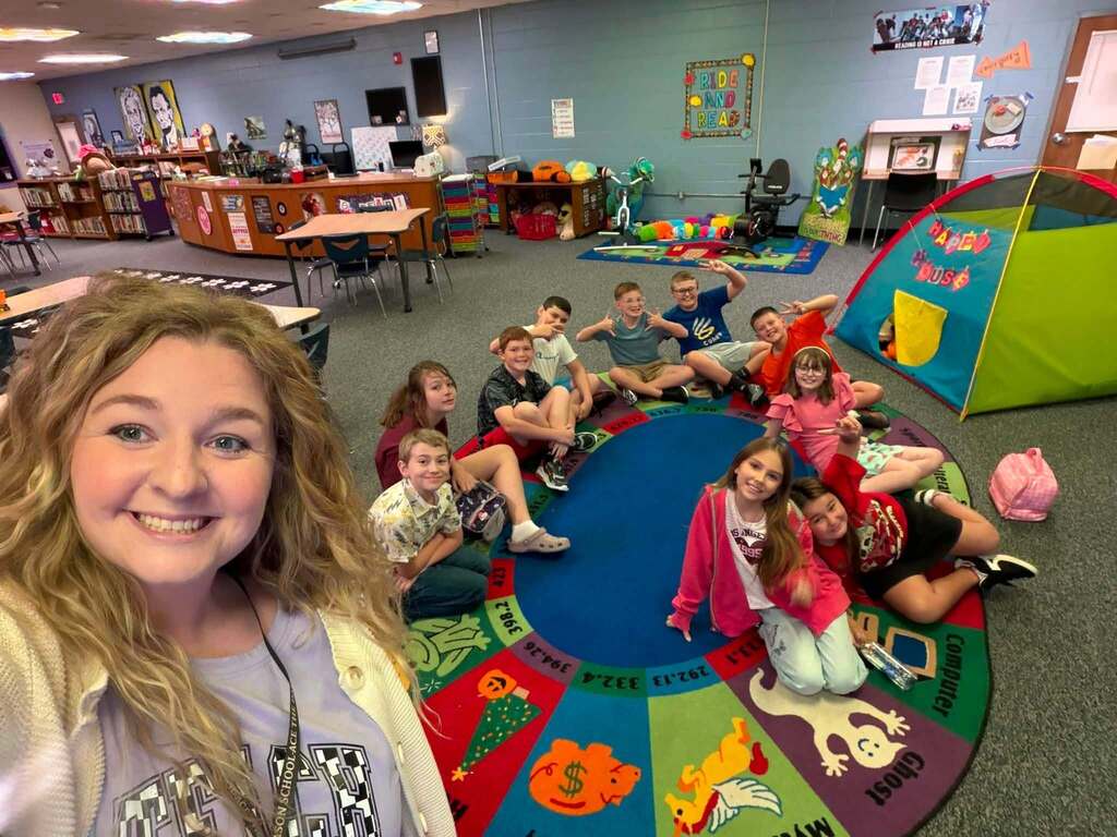 A woman stands smiling in front of a group of children sitting on a multicolored rug.