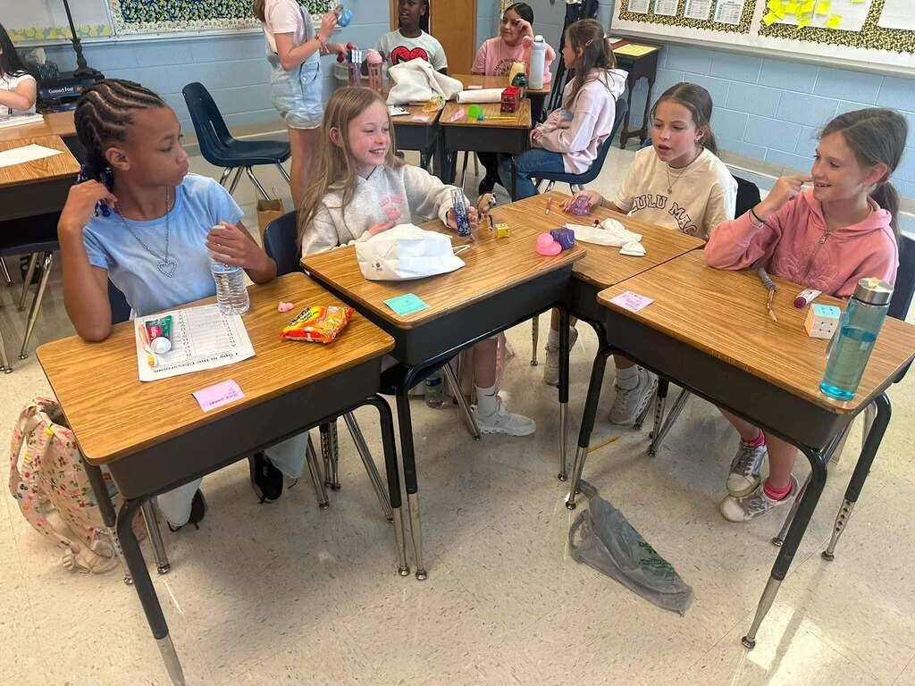 Four girls sit at desks in a classroom, each with papers and snacks, sharing and smiling.