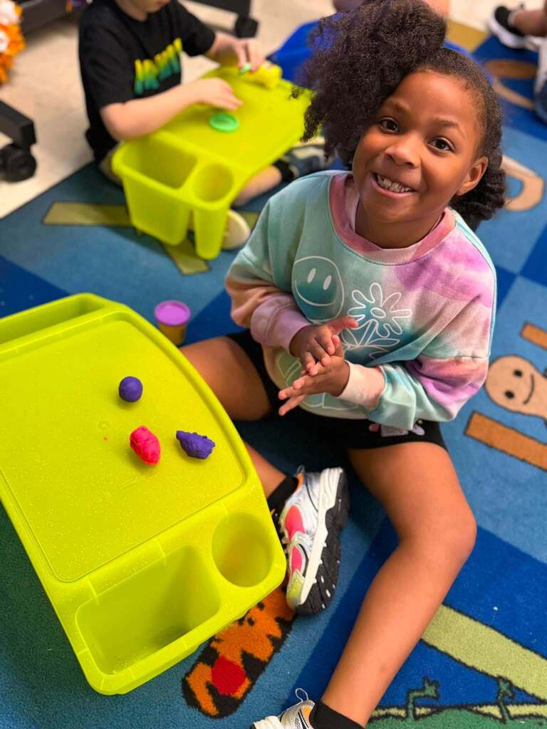 Two children seated on a floor, playing with a toy tray with small objects, one smiling.