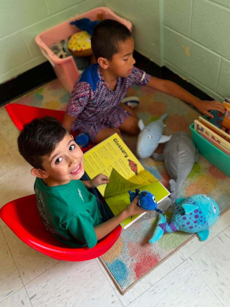 Two children sit on chairs, one reading a book and the other holding a toy. A rug, toys, and storage boxes are on the floor.