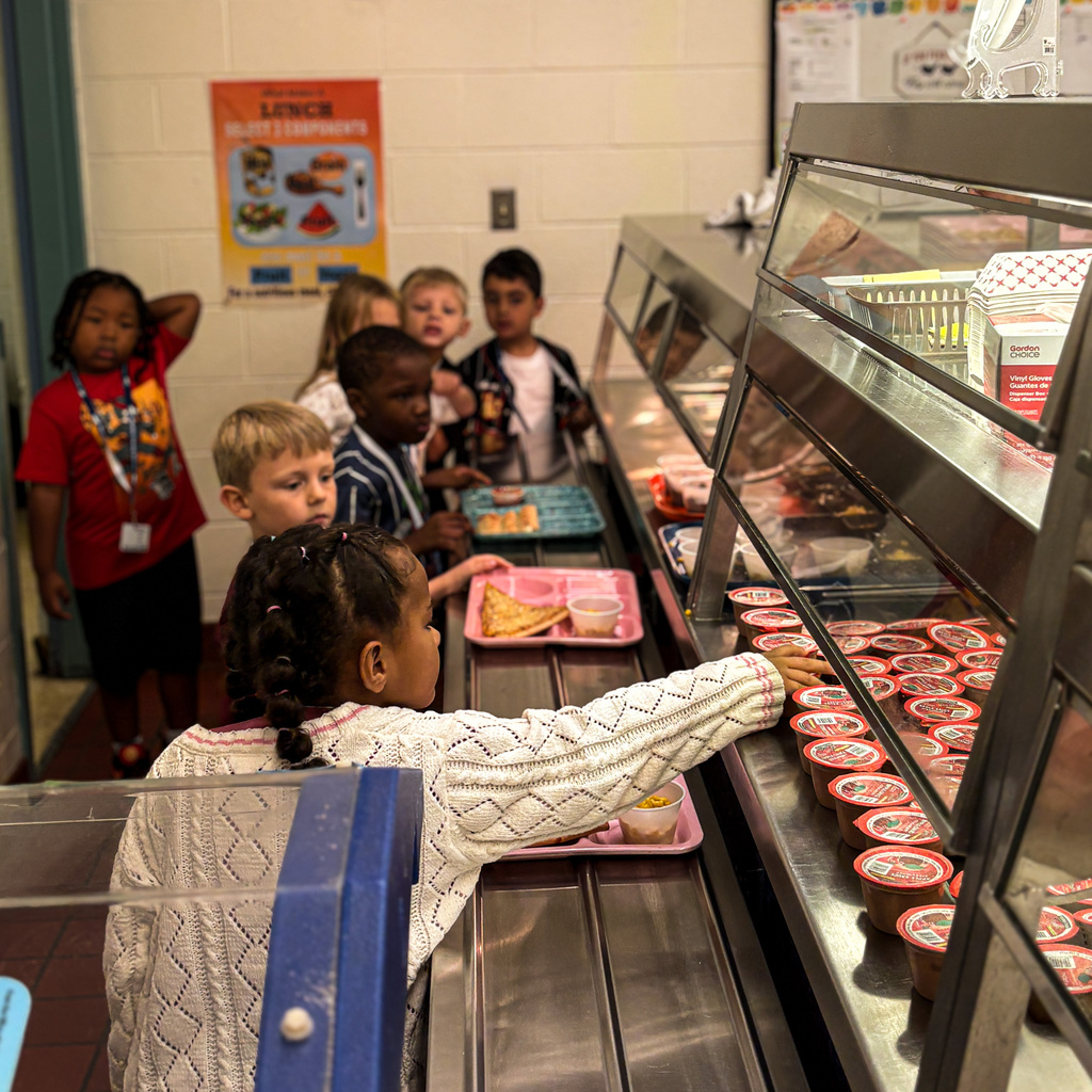 Children at a cafeteria line, picking up food. A sign on the wall in the background.