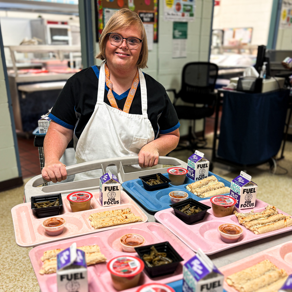 A smiling woman with glasses and an apron stands behind a table with trays of food.