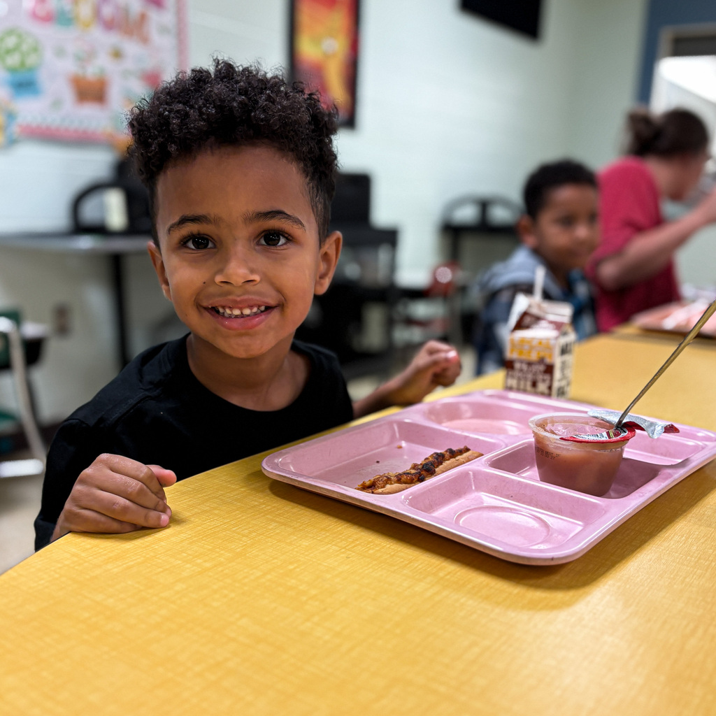 A boy with curly hair sits at a school table with a tray of food and a drink.