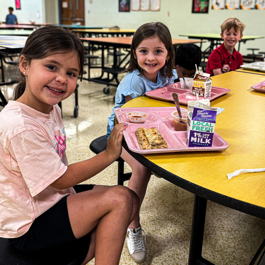 Children sit at a round table, smiling. Trays hold food and milk. Walls have artwork. Tables and chairs are arranged in a room.