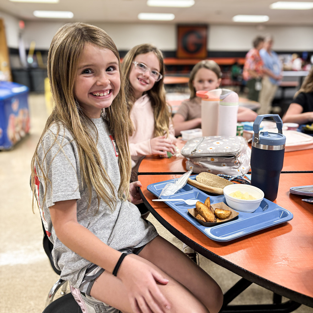 A girl in a gray shirt sits at a table in a cafeteria, smiling and looking at the camera.