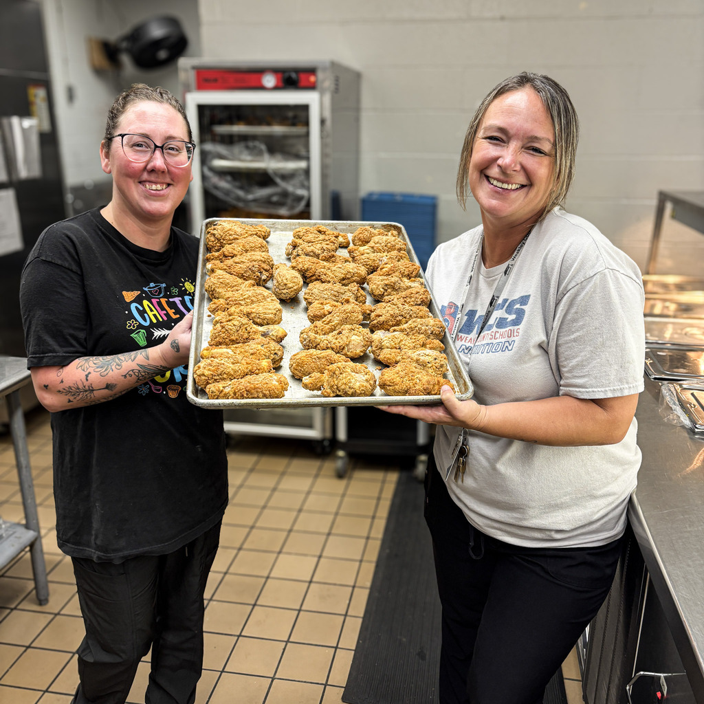 Two women hold a baking tray filled with chicken drumsticks. One woman wears glasses. They stand in a kitchen.
