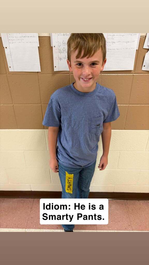 A smiling boy stands against a wall with white papers and brown tiles. He wears a blue shirt and jeans. Text reads, "Idiom: He is a Smarty Pants."