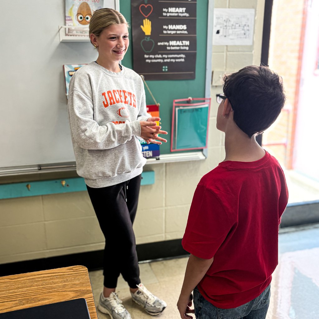 Two students in a classroom; a girl and boy are standing facing each other, conversing.
