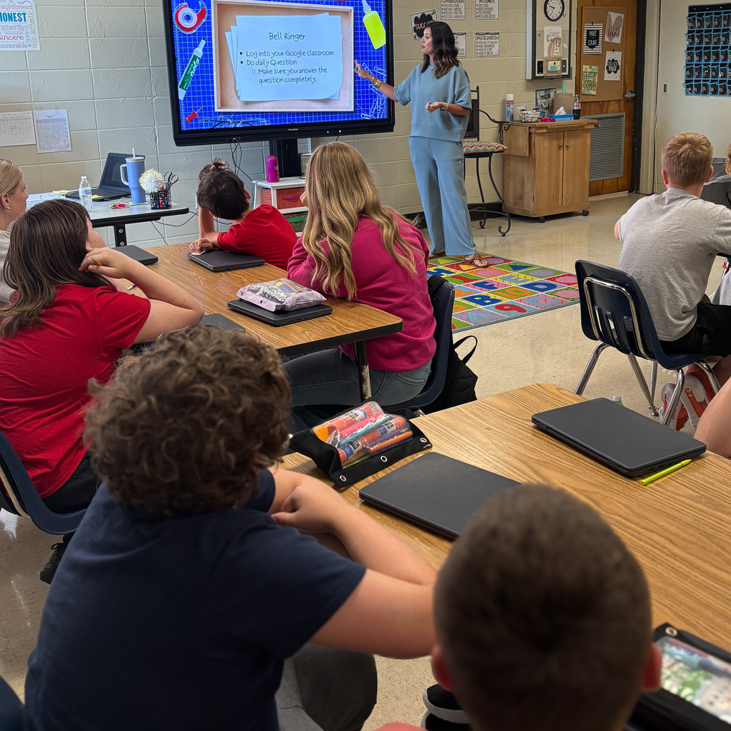 Students sit at desks in a classroom, facing a teacher who stands before a screen with a presentation.