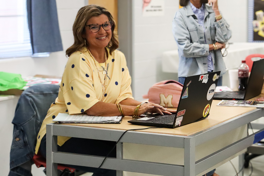 A smiling woman in a yellow polka-dot top sits at a desk typing on a laptop covered in university and school stickers during a staff training session.