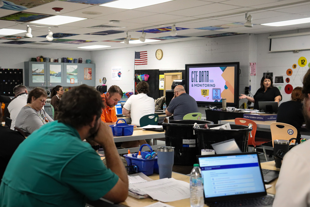 A room full of teachers and staff sit at desks with laptops during a CTE Data and Monitoring training session, with a presenter speaking at the front near a large display screen.