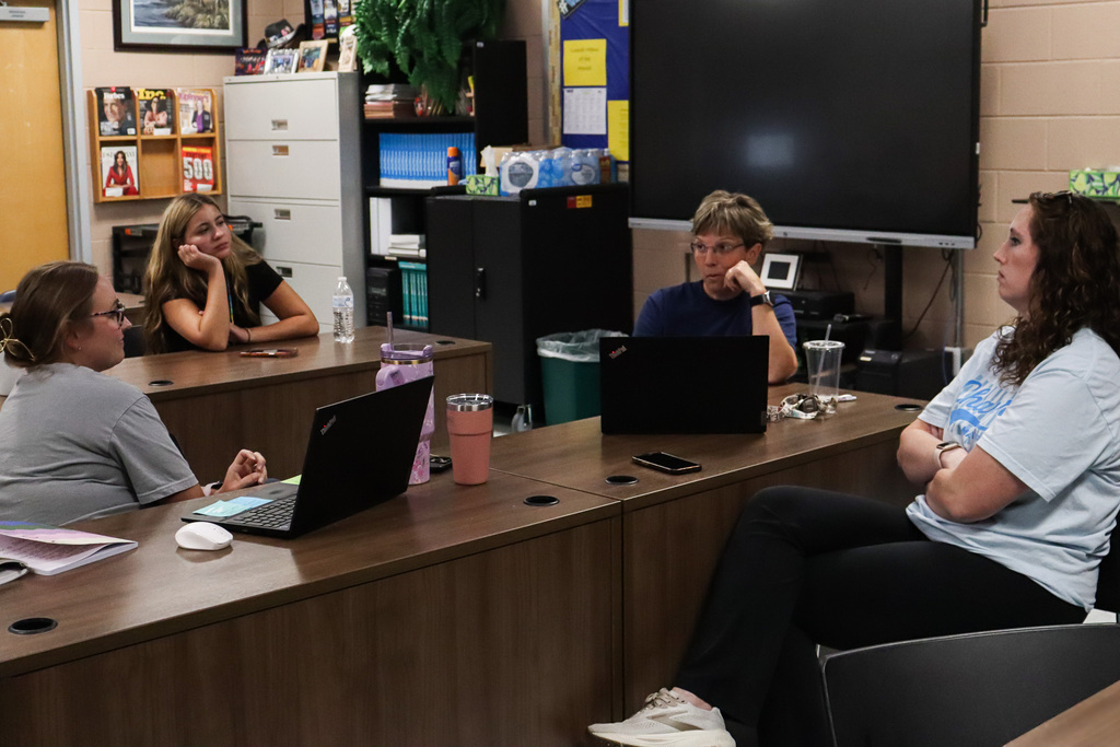 Four staff members sit around connected desks with laptops open, engaged in conversation during a small-group training session in a classroom.