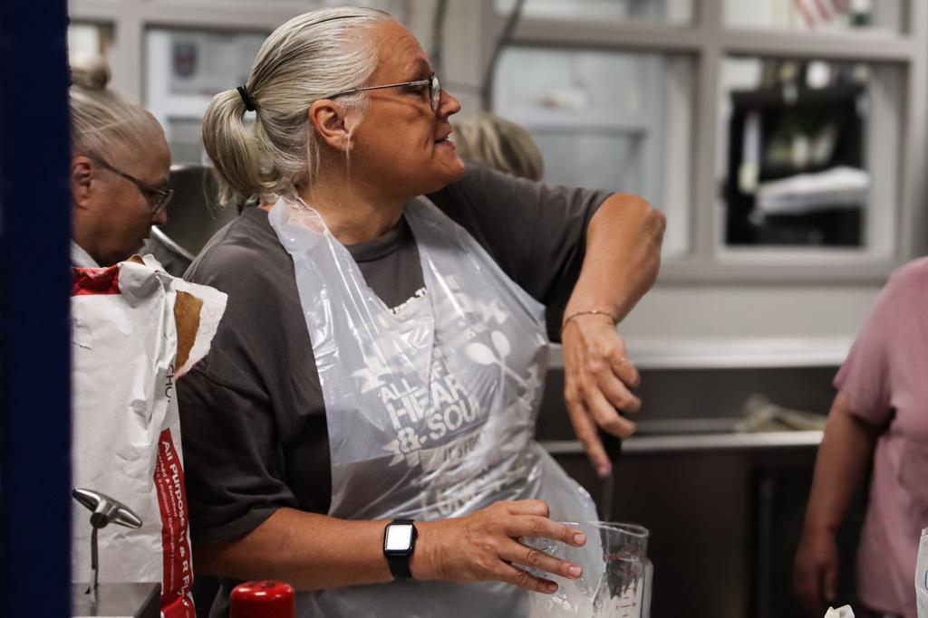 A cafeteria staff member with a gray ponytail and glasses, wearing a clear plastic apron, reaches for a mixer while glancing over her shoulder and smiling during a food prep training session.