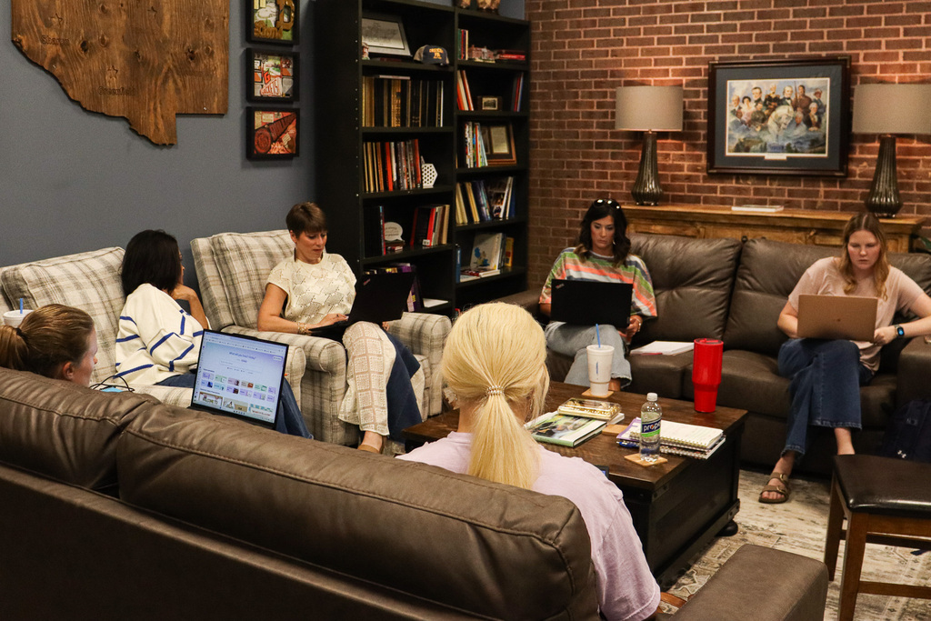 A group of staff members sit on couches and chairs with laptops open, collaborating in a cozy lounge space decorated with school memorabilia and a wooden map of Weakley County communities.