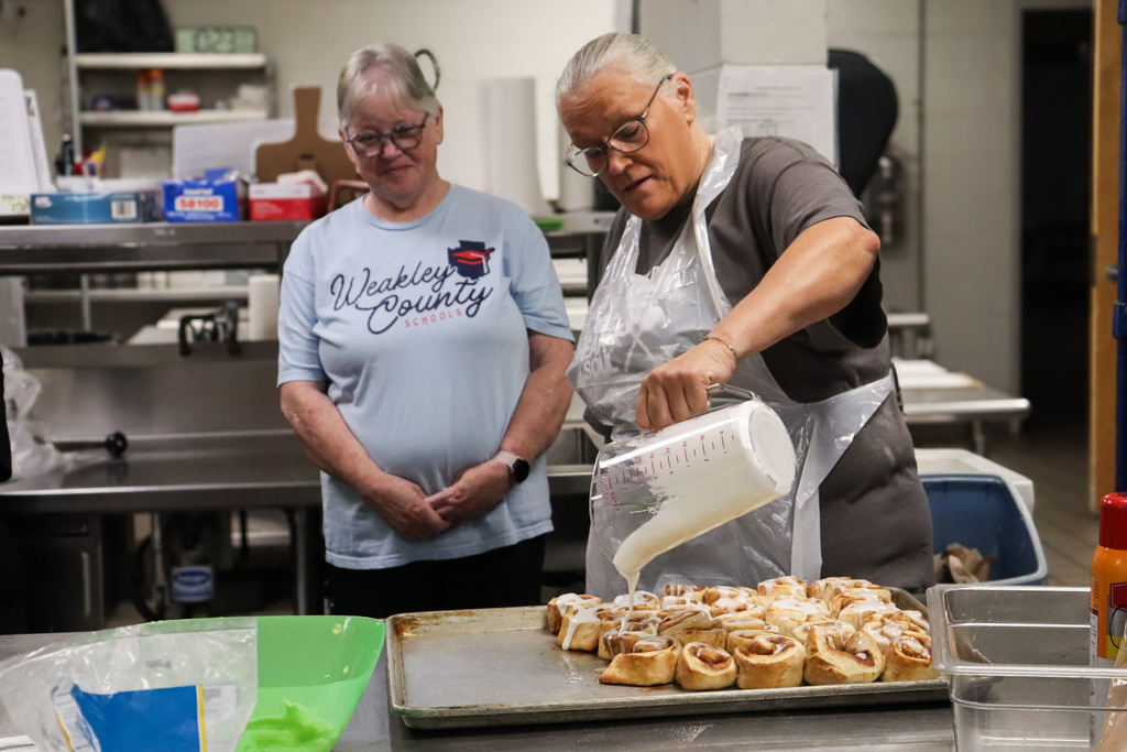 A cafeteria worker in a Weakley County Schools t-shirt watches as a colleague in a plastic apron pours icing over a tray of fresh cinnamon rolls.