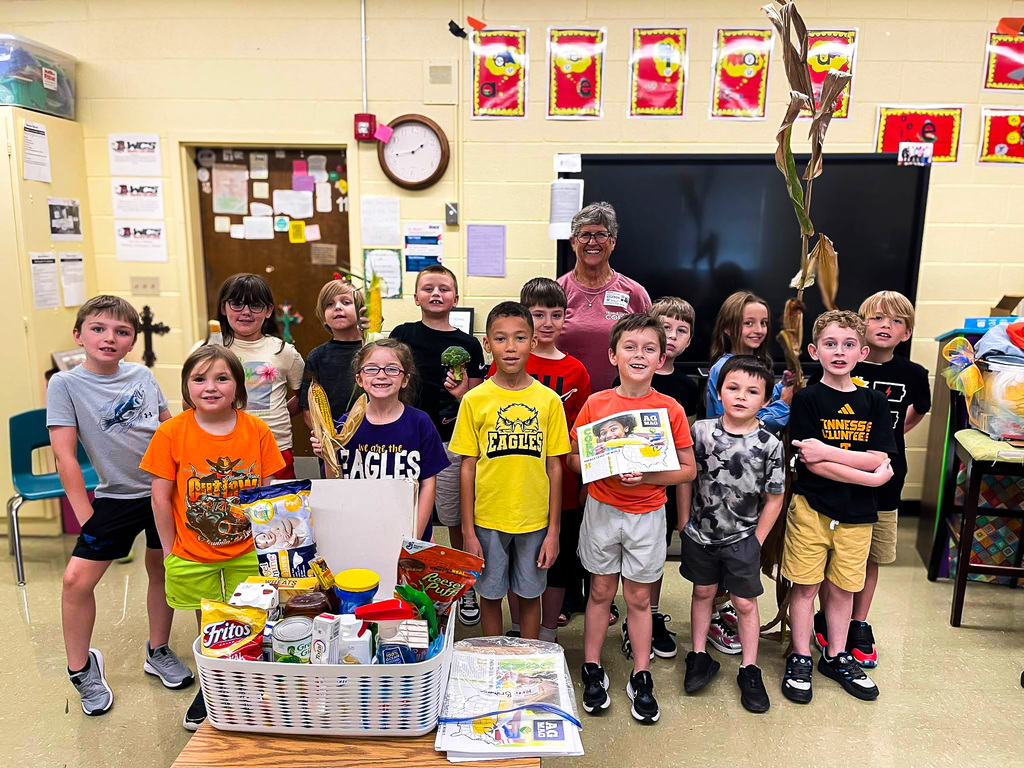 Miss Terri Brundige stands with a class of elementary students following their lesson about corn. Students hold ears and stalks of corn and examples of everyday products that contain corn, with a basket of products displayed in front of the group.
