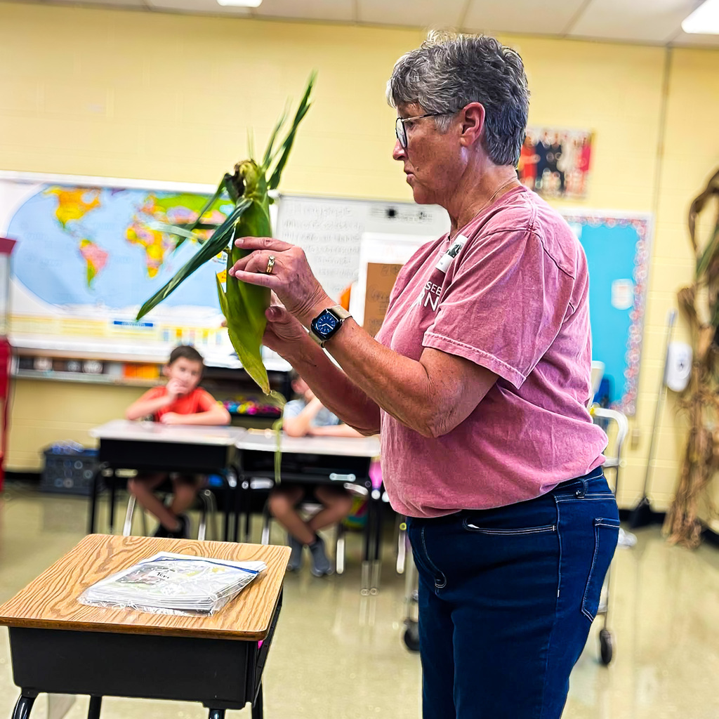 Miss Terri Brundige holds an ear of corn still in its husk as she talks with elementary students about how corn grows and is harvested.