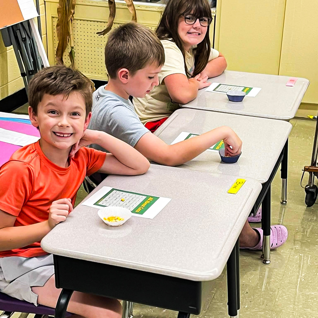 Three elementary students sit at their desks playing Corn Bingo, using corn kernels as markers on their bingo cards. One student smiles toward the camera while the others work on their cards.