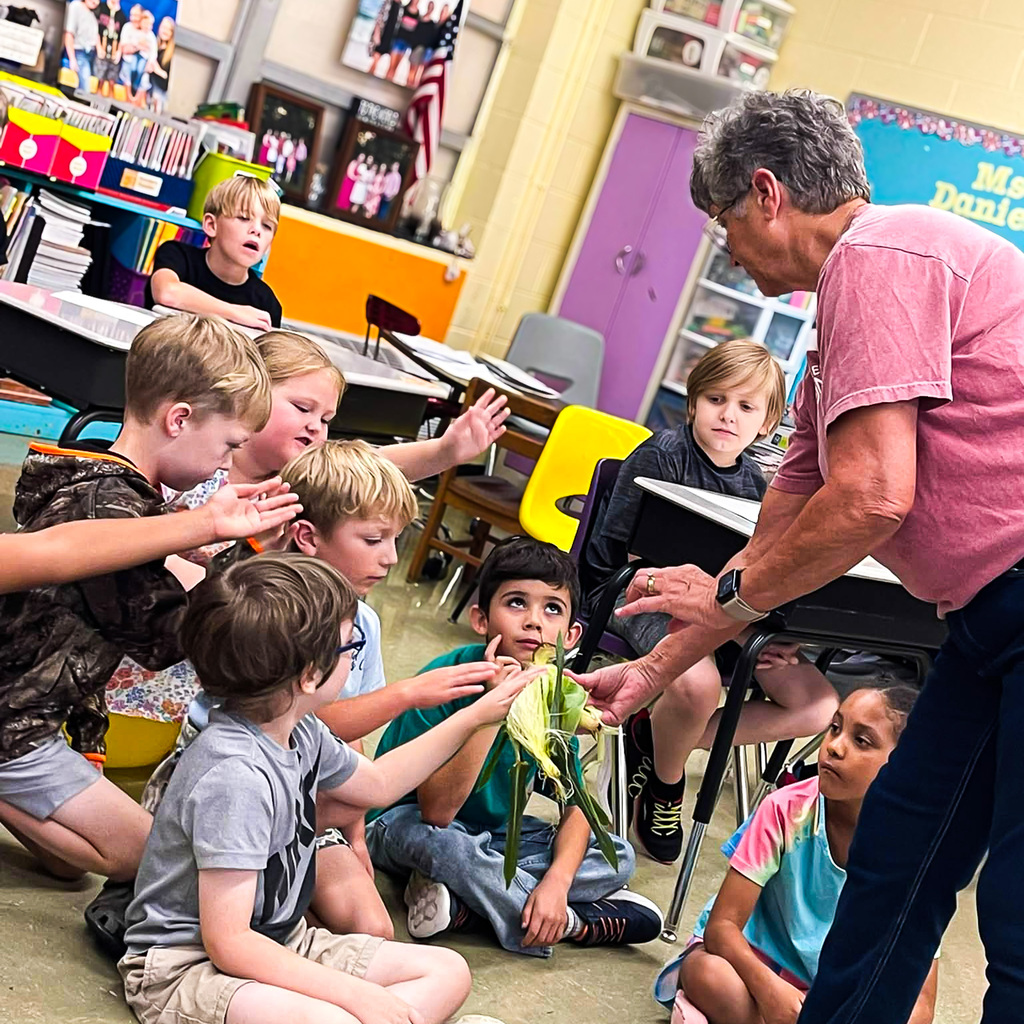 A group of elementary students gathers around Miss Terri Brundige to get a closer look at a partially husked ear of corn, with several students reaching out to touch the husk and silk.