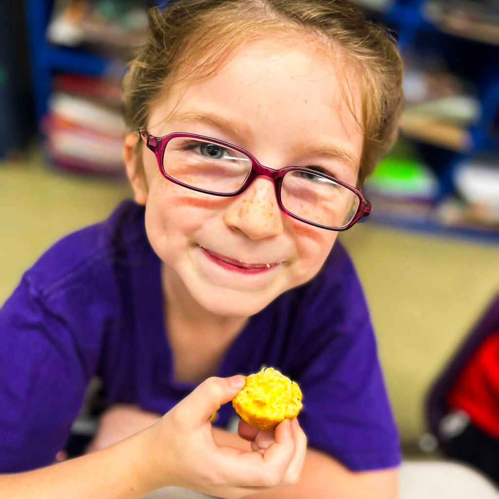 A smiling elementary student wearing glasses holds up a small corn muffin after a classroom lesson about corn.