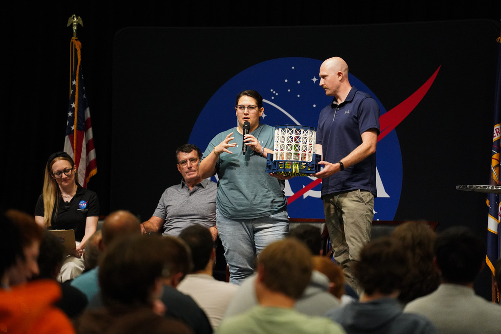 NASA professionals demonstrate a model for students during a CTE Day presentation, with a large NASA logo displayed behind them.