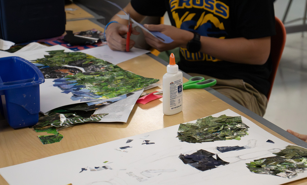 A Westview student works on a magazine mosaic at a classroom table. Small pieces of green and blue magazine pages are arranged over a sketched design, with scissors, glue, and additional magazine pages nearby.