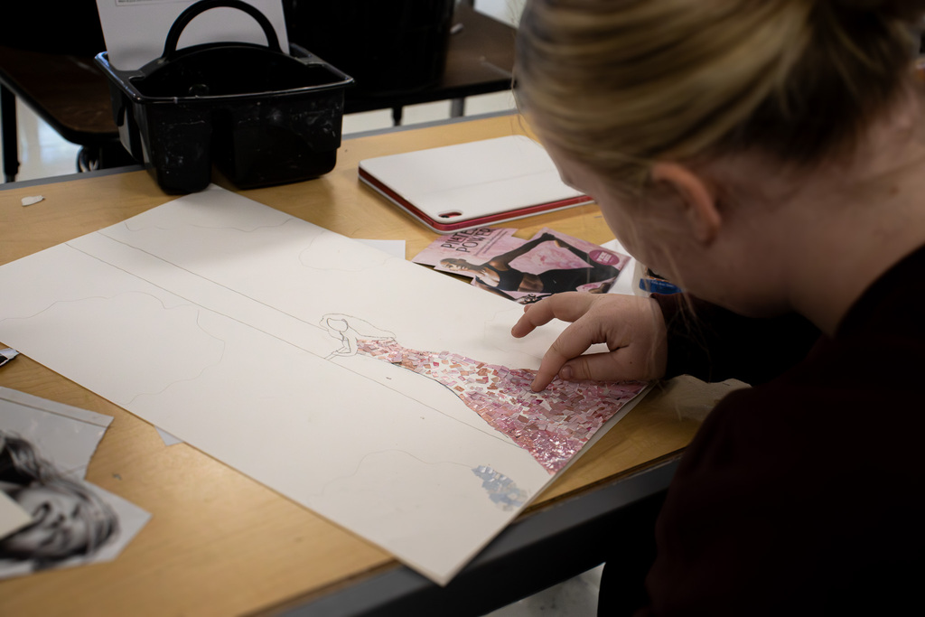 A Westview student carefully places small pink magazine pieces onto a sketched figure wearing a long dress, building the mosaic section piece by piece.