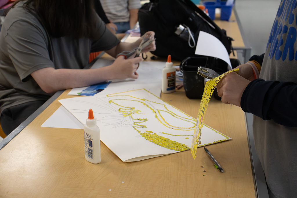 Two Westview art students cut pieces from magazine pages for their mosaic projects. A partially completed yellow mosaic design, scissors, glue, and paper scraps cover the worktable.