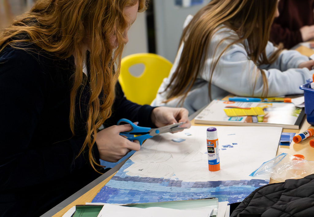 A Westview student uses scissors to cut small blue pieces for a mosaic artwork. Rows of blue magazine pieces have already been glued along the bottom of the sketched design.