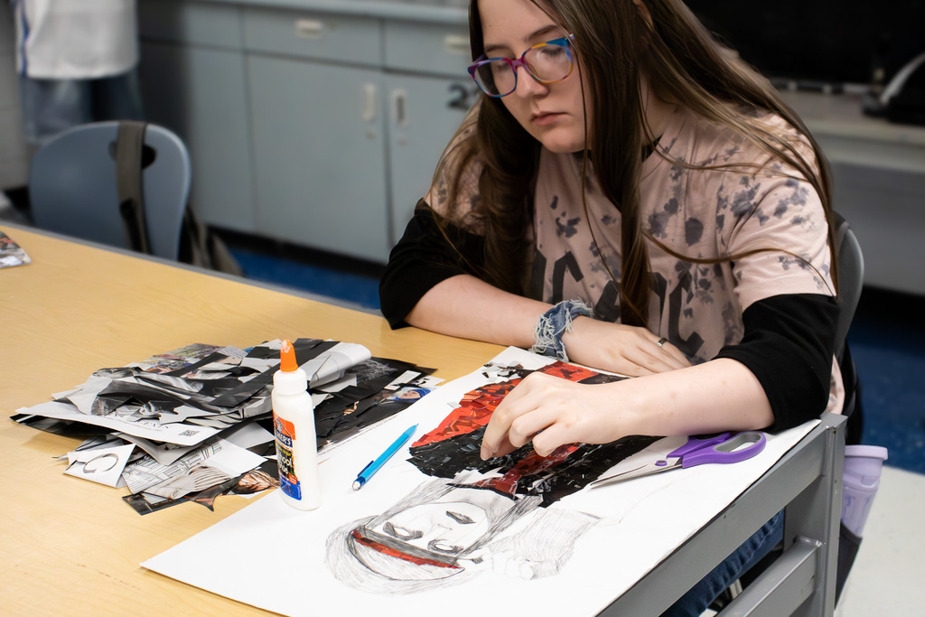 A Westview art student works on a large portrait mosaic, placing red and black magazine pieces over a sketched figure. Cut magazine pages, scissors, glue, and a pencil surround the artwork.
