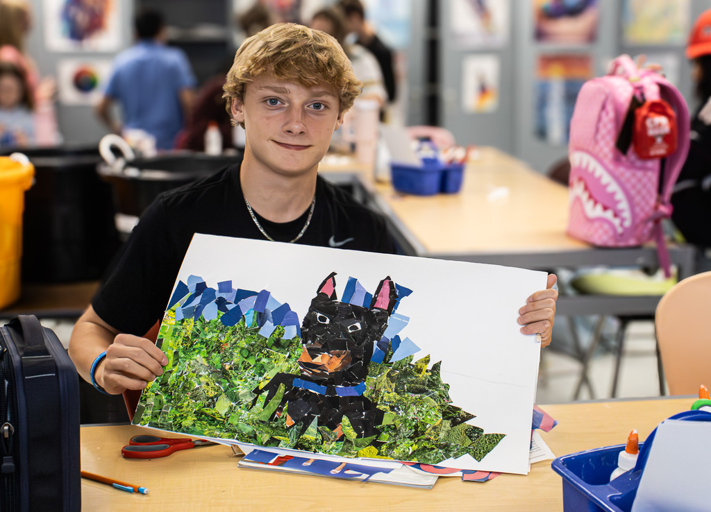A Westview student smiles while holding up his in-progress magazine mosaic of a black dog sitting in a green landscape with blue mountains. The image is constructed from colorful pieces cut from magazine pages.