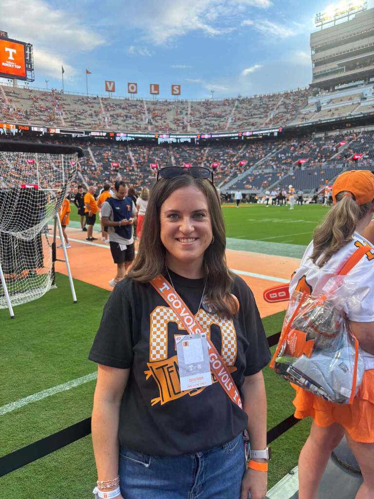 Sharon School teacher Caitlin Tucker smiles on the field at Neyland Stadium before a Tennessee football game. She wears a Tennessee-themed T-shirt and game credential, with the football field and stadium seating visible behind her.