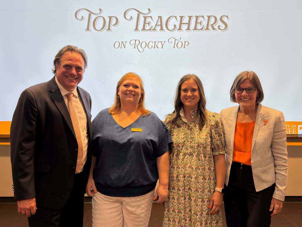 Four people stand together and smile for a photo in front of a screen reading “Top Teachers on Rocky Top” at the University of Tennessee. Sharon School teacher Caitlin Tucker stands second from the right in a floral dress.