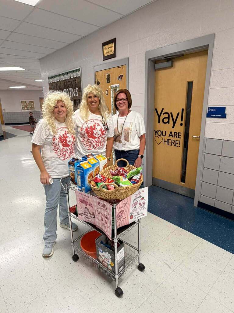 Three school staff members celebrate Dolly Day in a hallway, including two wearing blonde Dolly-inspired wigs. They stand beside a decorated snack cart featuring Dolly Parton-themed signs.