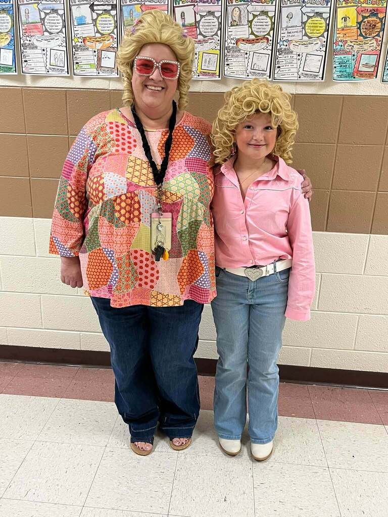 A staff member and student pose together in Dolly-inspired outfits, both wearing curly blonde wigs. The student wears a pink shirt, jeans, a rhinestone belt, and white boots.