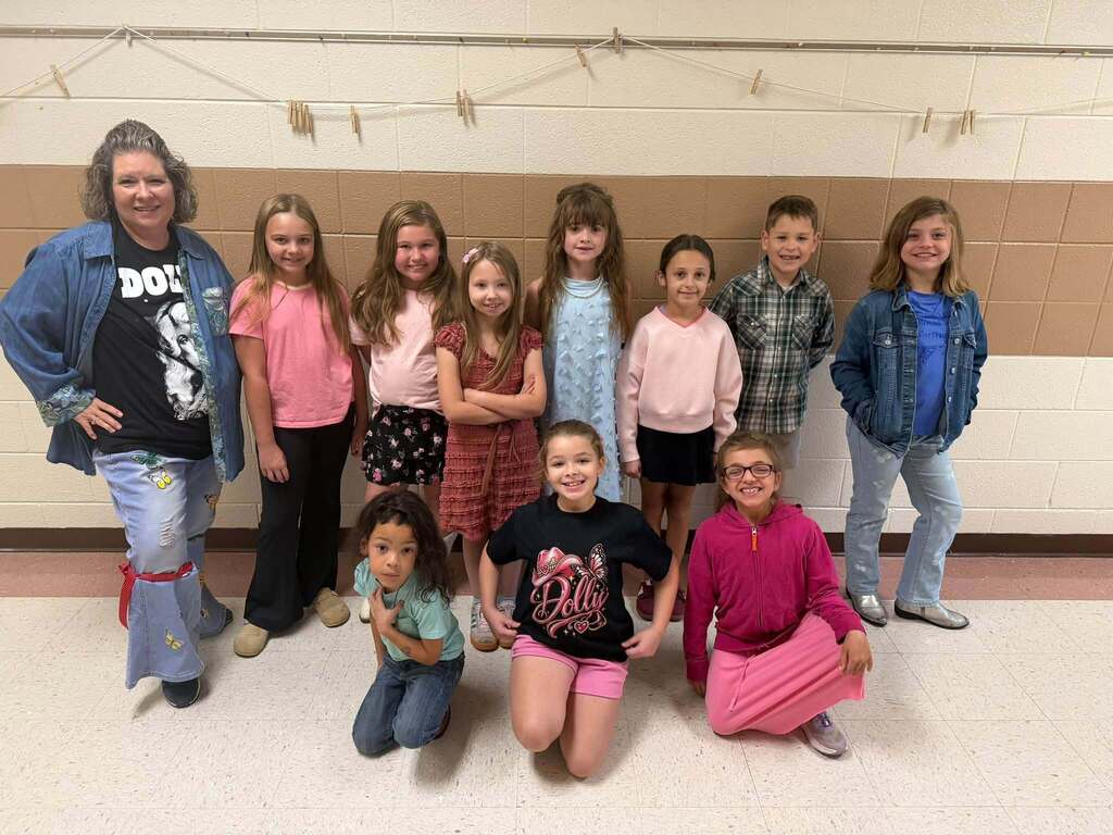 A staff member poses with a group of elementary students celebrating Dolly Day. Students wear pink, denim, Dolly Parton shirts, and other Dolly-inspired clothing.