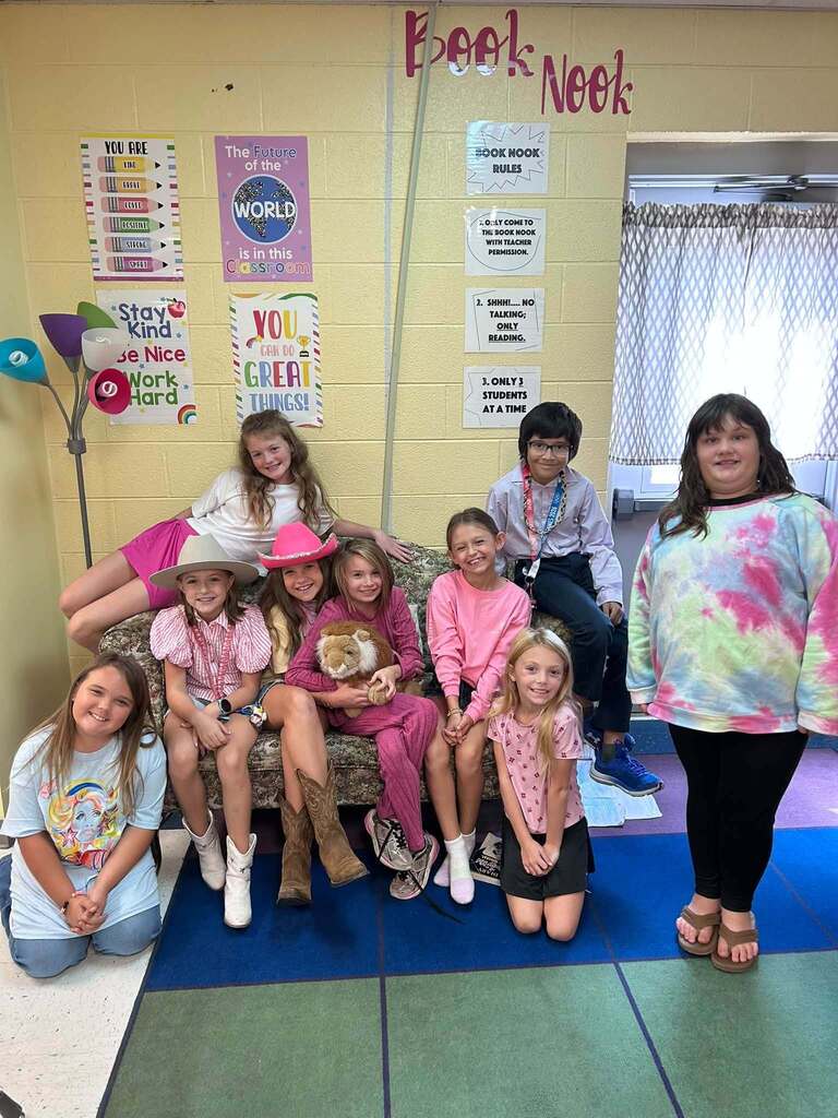 A group of elementary students pose together in a classroom book nook while celebrating Dolly Day. Several students wear pink, cowboy hats, boots, and other Dolly-inspired clothing.